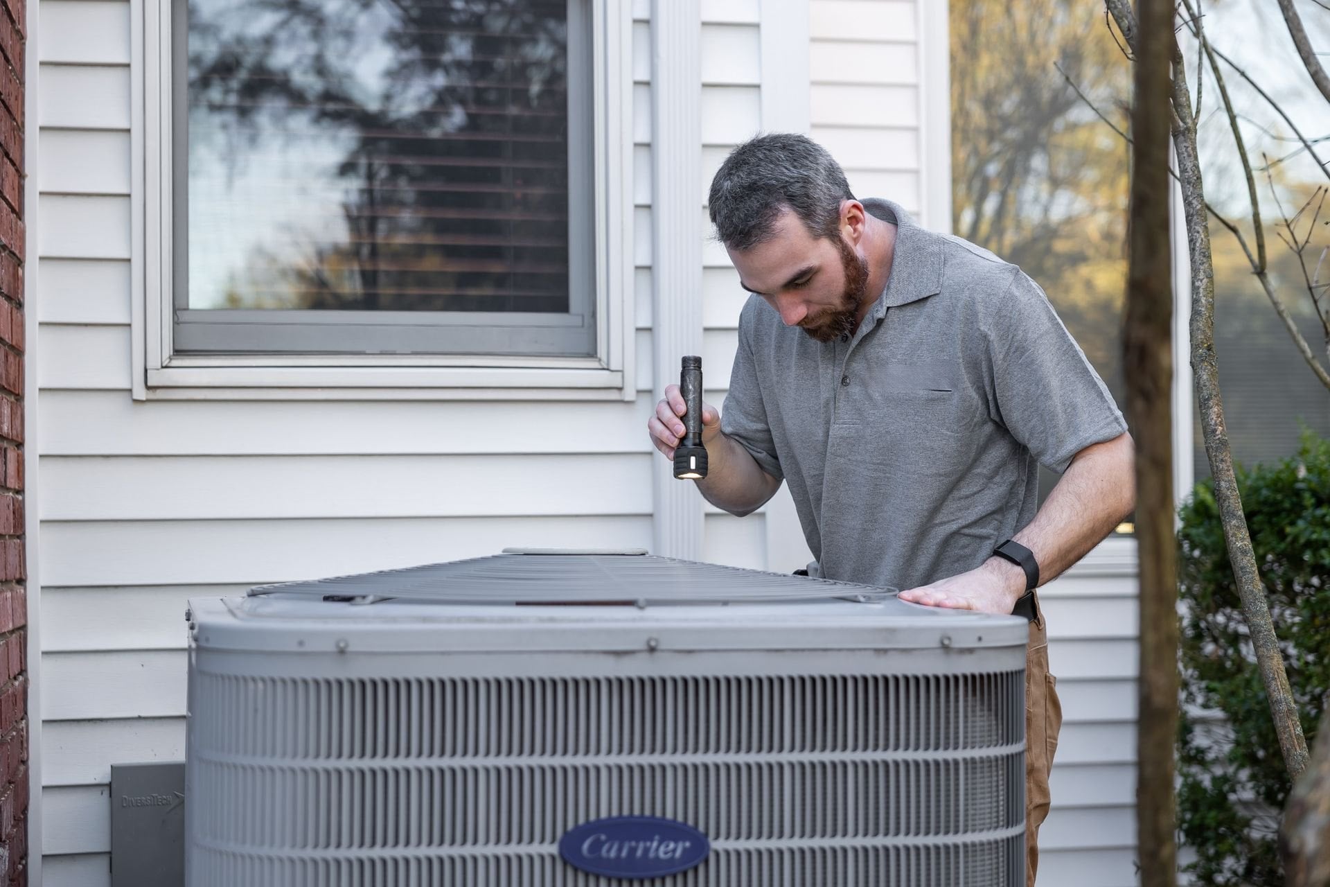 An HVAC technician with a flashlight inspecting an outdoor air conditioning condenser beside a house
